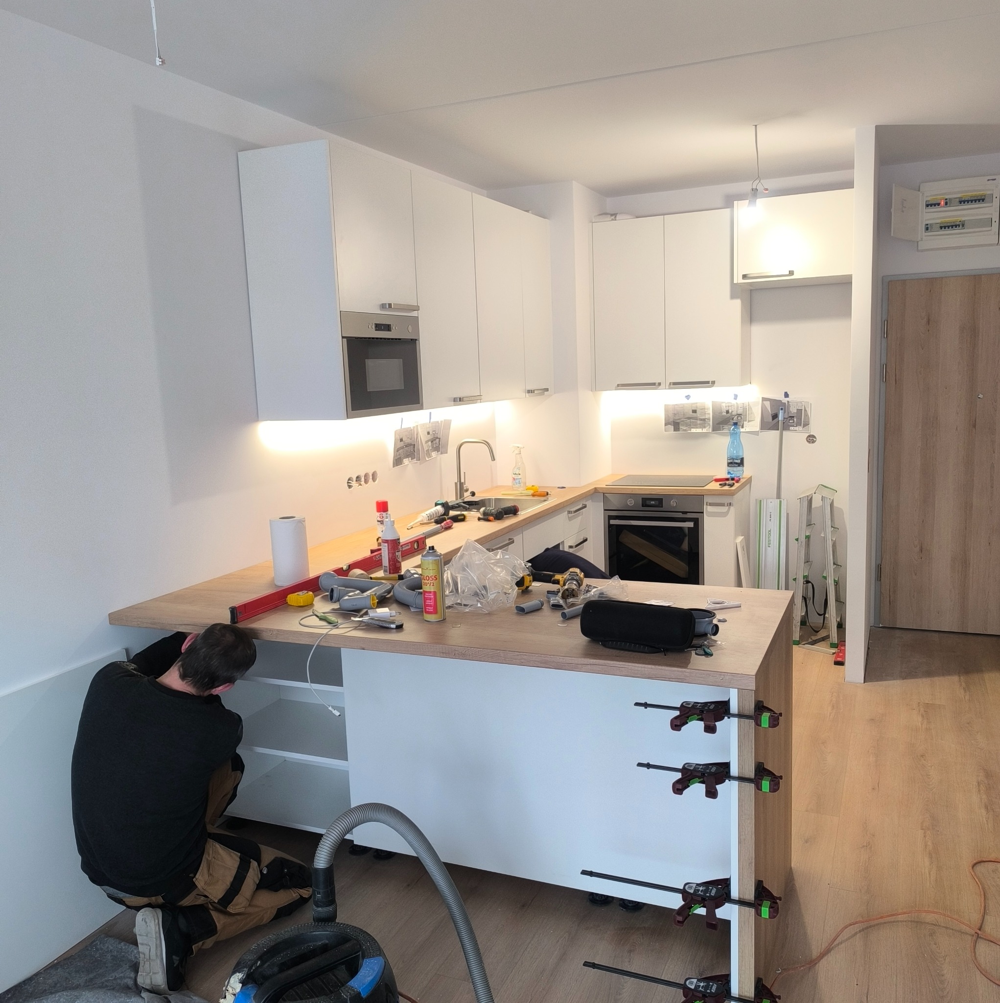 Montaż blatu kuchennego w Swarzędzu: stolarz instaluje drewniany blat, widoczne narzędzia, poziomica, klamry mocujące i odkurzacz, białe szafki kuchenne w tle.