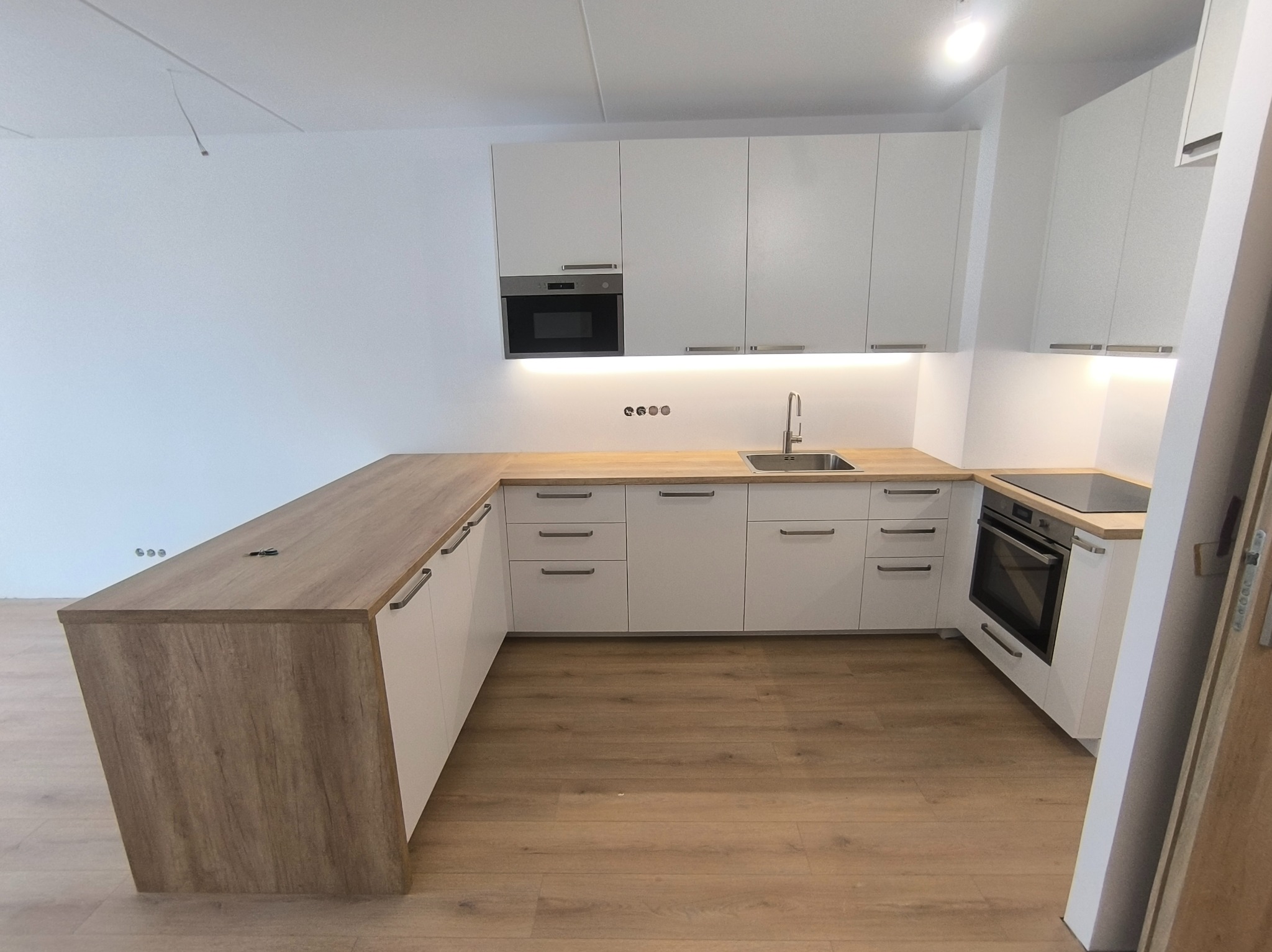 Nowoczesna, minimalistyczna kuchnia z białymi szafkami i drewnianym blatem, oświetleniem LED pod szafkami wiszącymi i drewnianą podłogą, widziana z niskiego kąta.