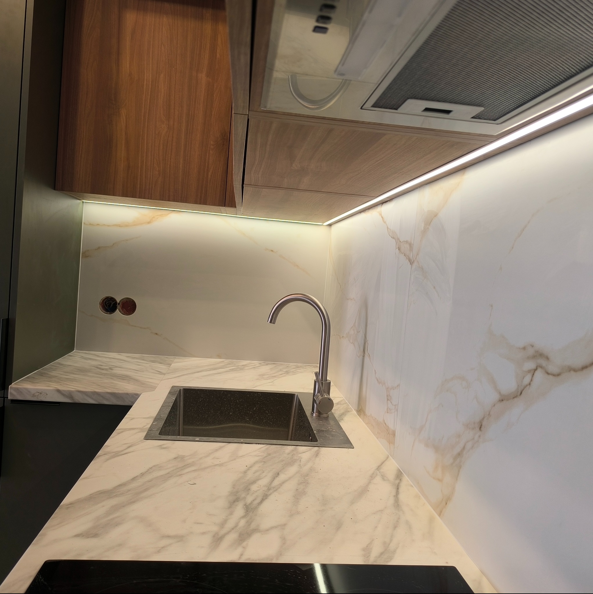 Nowoczesna kuchnia z marmurowym blatem i ścianą, oświetleniem LED pod szafkami i metalowym zlewem ze stali nierdzewnej.