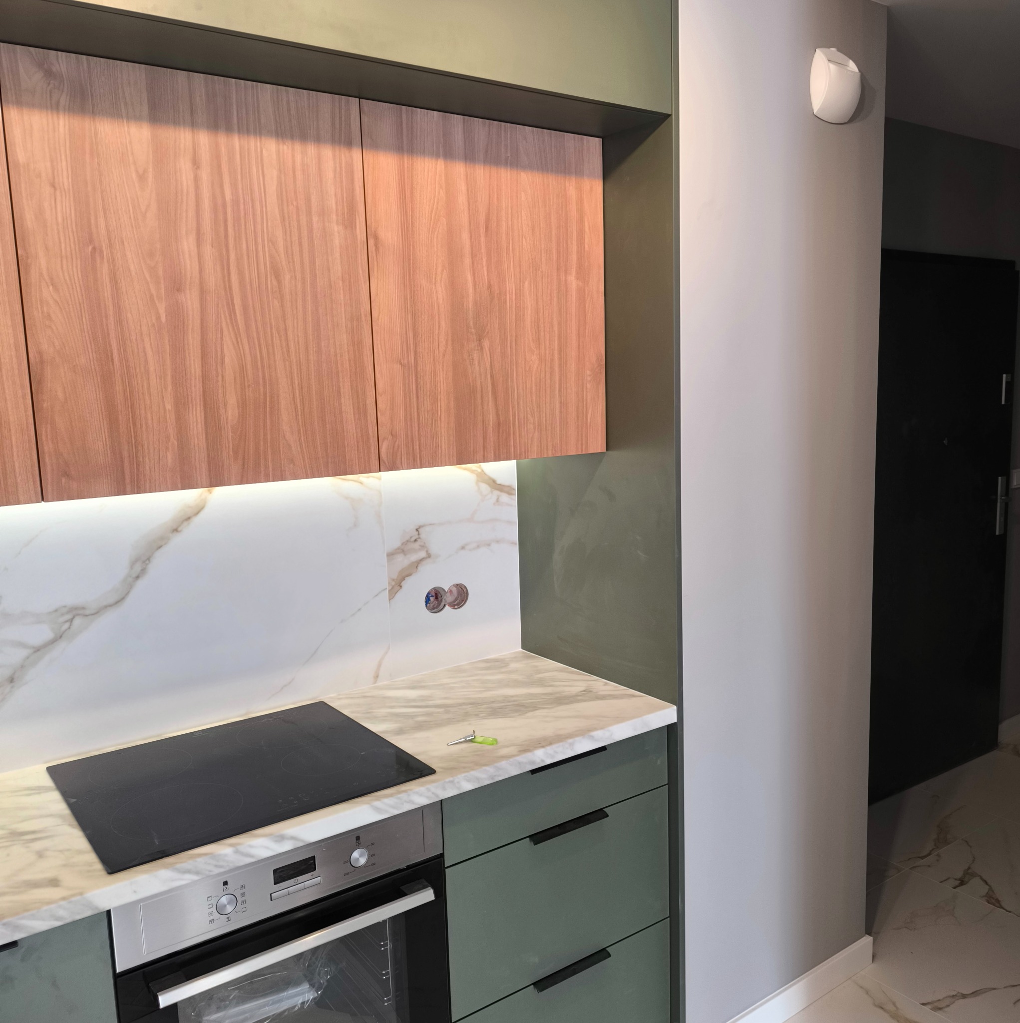 Fragment kuchni z drewnianymi szafkami górnymi, blatem z imitacją marmuru, płytą indukcyjną i zielonymi szafkami dolnymi. Na blacie leżą klucze.