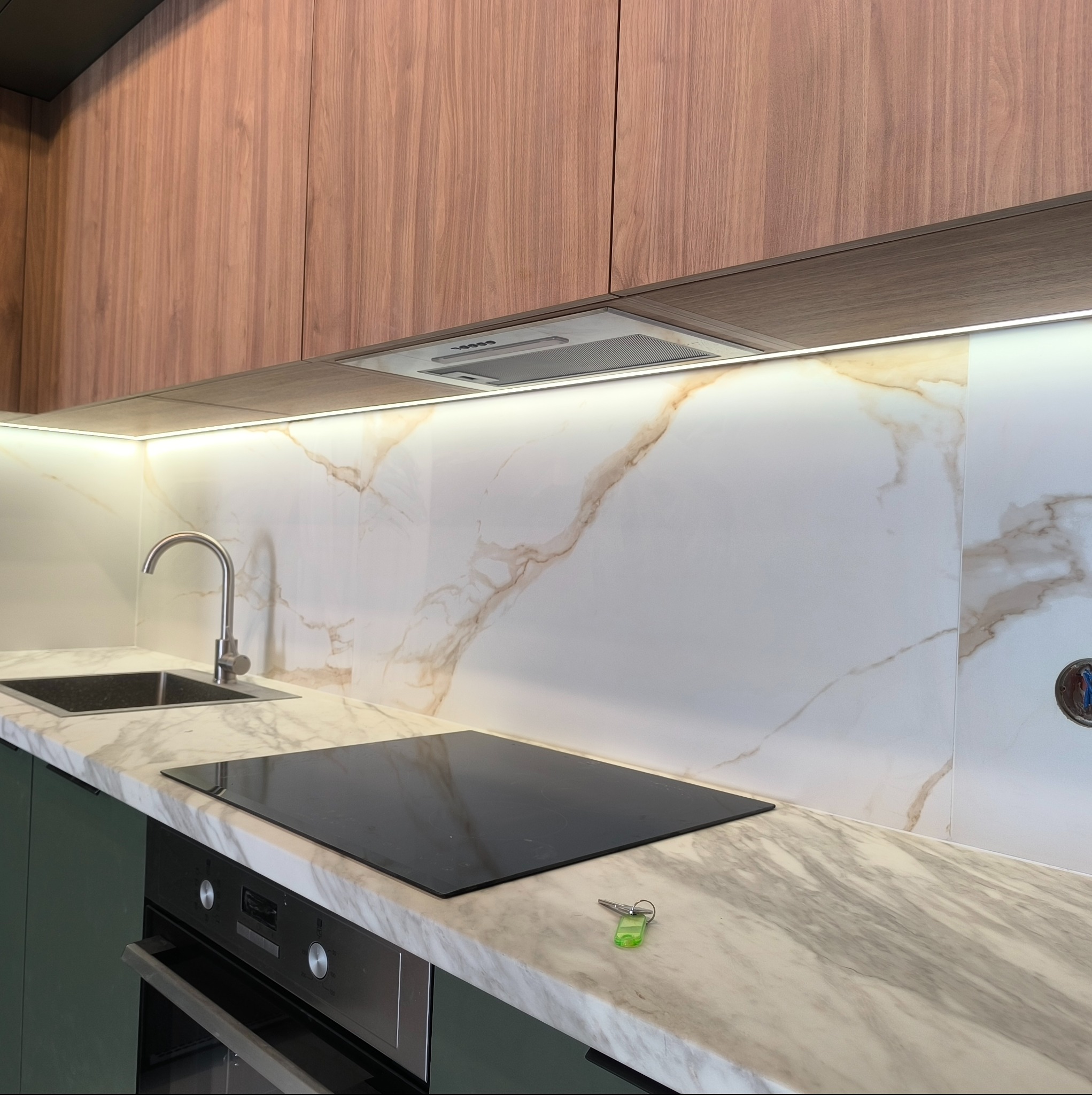 Nowoczesna kuchnia z drewnianymi szafkami górnymi, blatem imitującym marmur, płytą indukcyjną i zlewozmywakiem, oświetlona listwą LED pod szafkami, z kluczami leżącymi na blacie.