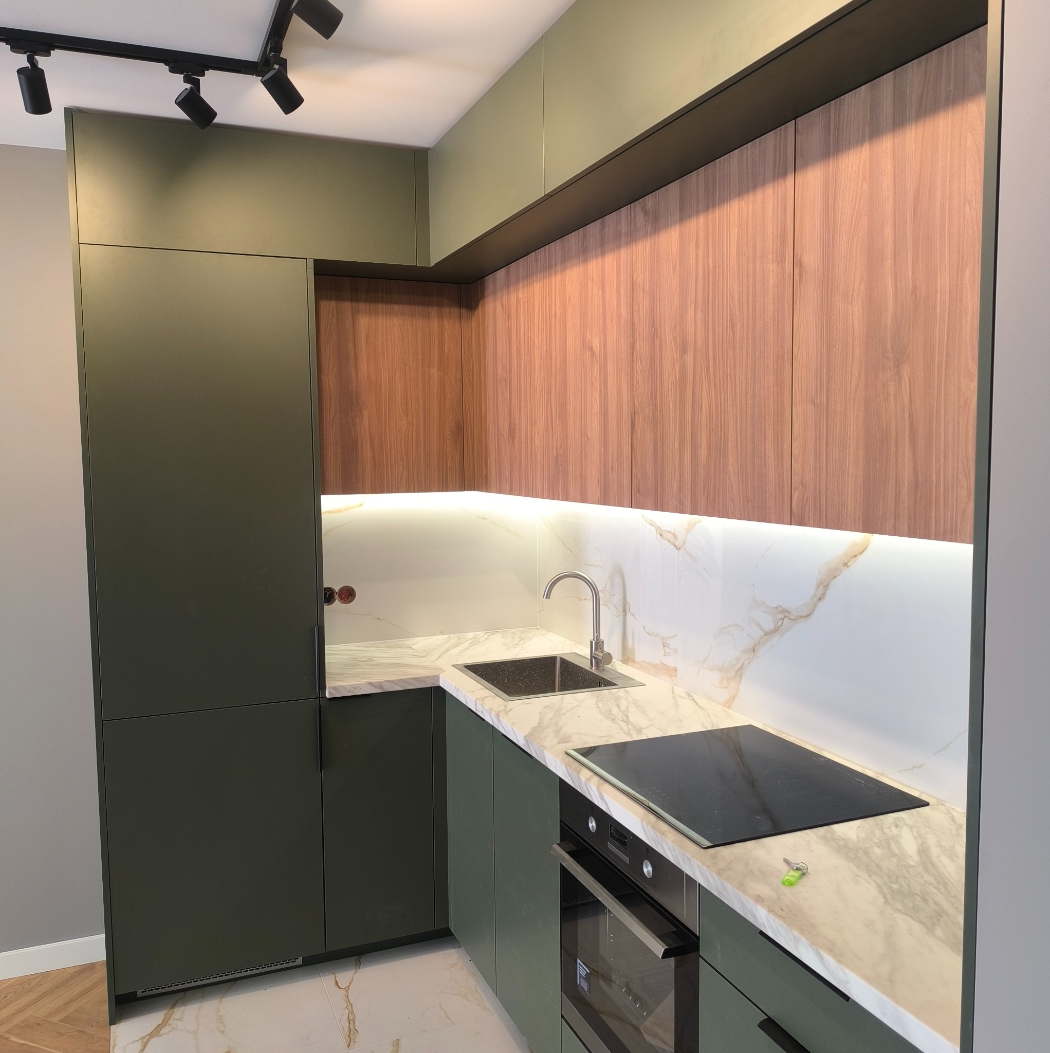 Nowoczesna kuchnia z oliwkowymi szafkami dolnymi i drewnianymi górnymi, marmurowy blat z płytą indukcyjną i zlewozmywakiem, oświetlenie LED pod szafkami, widoczne klucze na blacie.