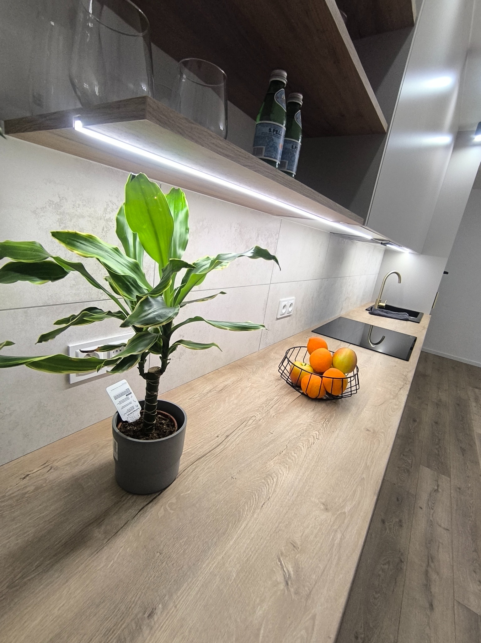 Jasny blat kuchenny z drewnianym wzorem, oświetlenie LED pod szafkami, roślina doniczkowa i koszyk z owocami, nowoczesny design.