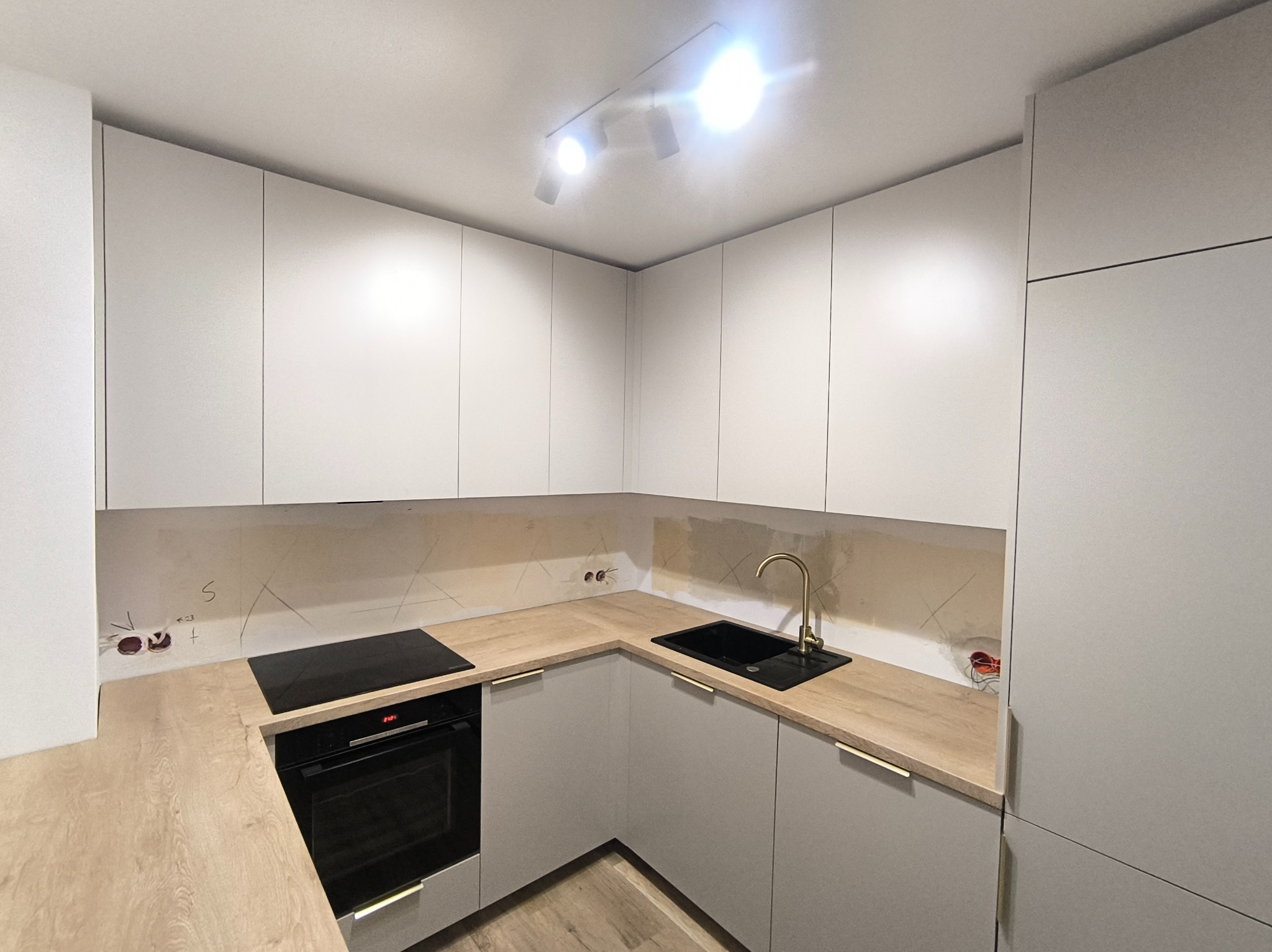 Wnętrze kuchni w trakcie montażu, widoczne szare szafki dolne i białe górne, drewniany blat, czarny zlew z złotą baterią, płyta indukcyjna, piekarnik oraz niedokończona ściana nad blatem...