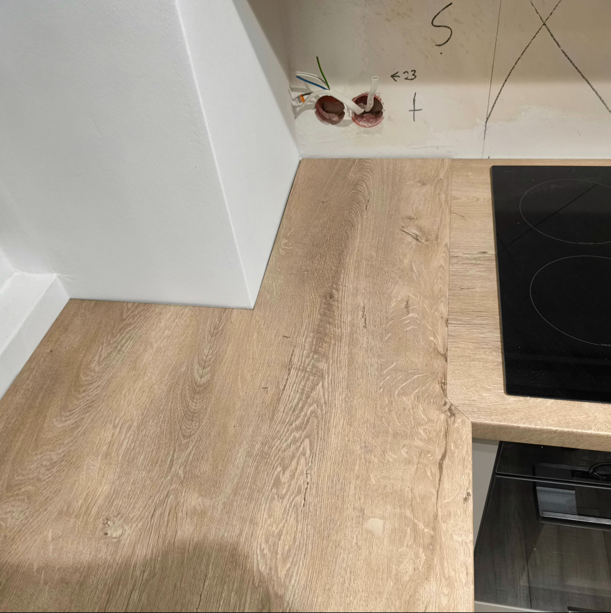 Fragment blatu kuchennego o jasnym, drewnopodobnym wzorze, przylegającego do białej ściany i płyty indukcyjnej; widoczne niedokończone gniazdka elektryczne i oznaczenia na ścianie.