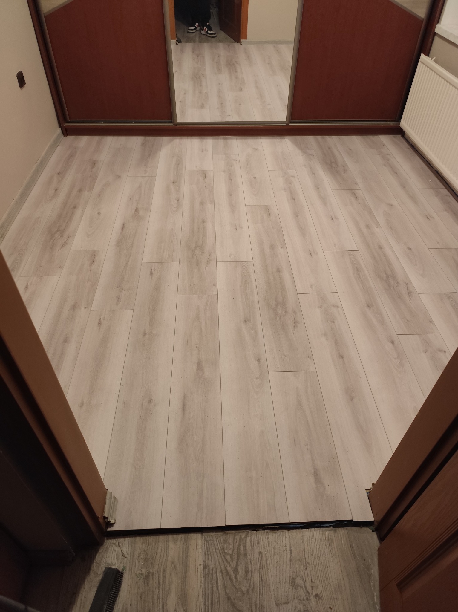 Jasne panele podłogowe ułożone wzdłuż korytarza, widoczne z perspektywy progu drzwi. W tle szafa z lustrzanymi drzwiami.