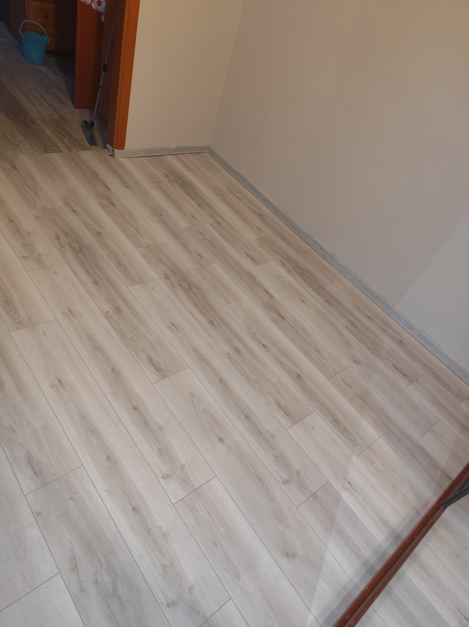 Jasne panele podłogowe ułożone w pokoju, widoczne listwy przypodłogowe i fragment progu drzwi. Neutralna kolorystyka ścian i podłogi.