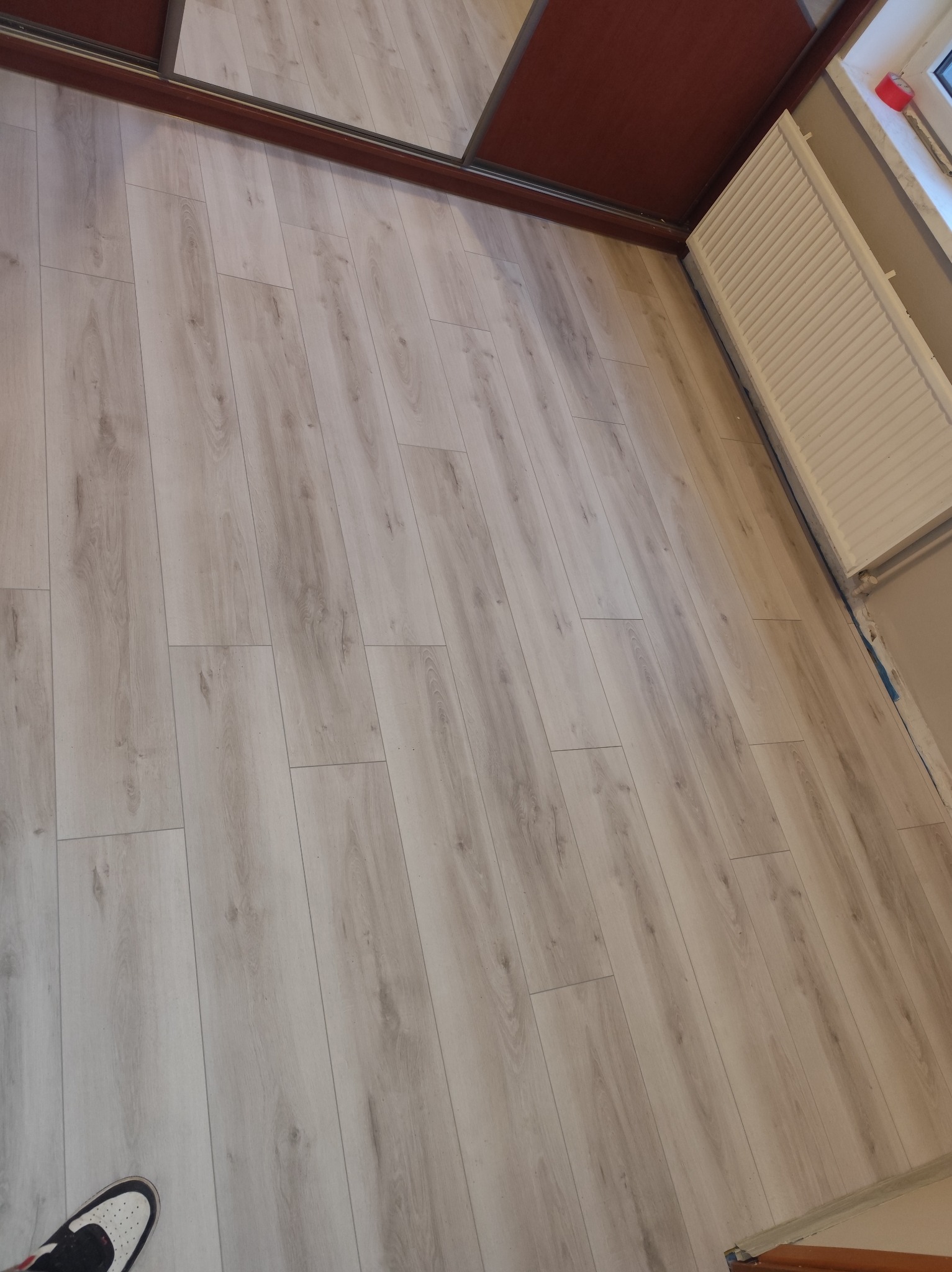 Jasne panele podłogowe ułożone wzdłuż ściany z grzejnikiem i brązowymi drzwiami przesuwnymi. Widoczny fragment buta w lewym dolnym rogu.