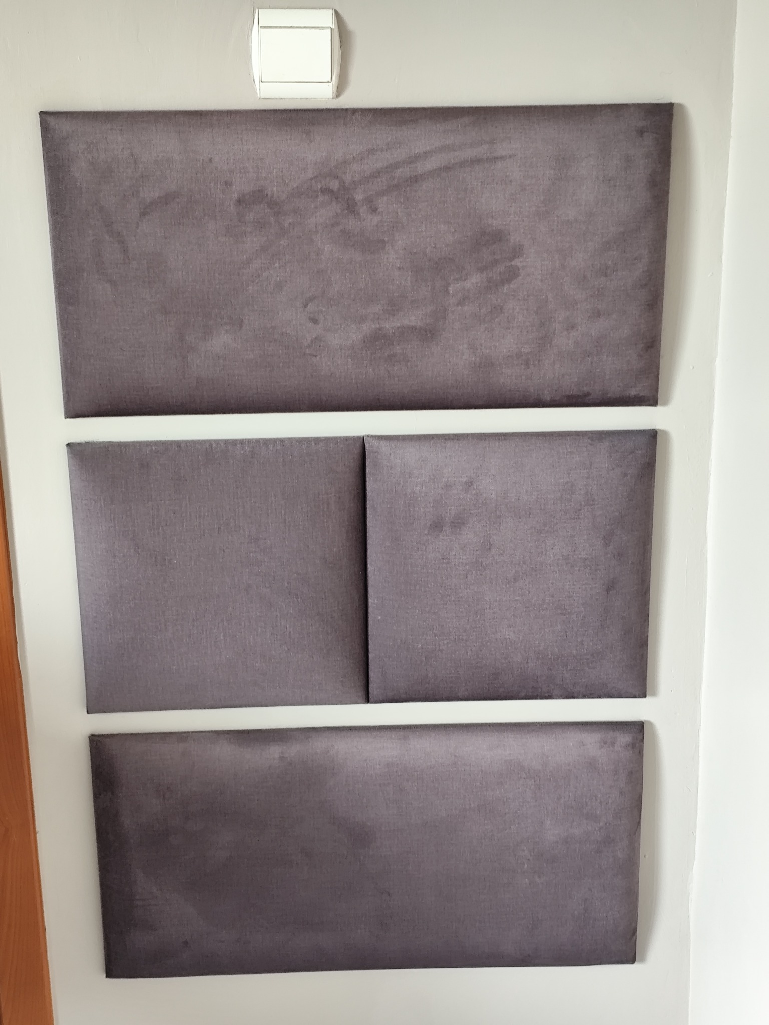 Zamontowane, szare panele tapicerowane na ścianie, tworzące dekoracyjny zagłówek. Widoczny włącznik światła nad górnym panelem.