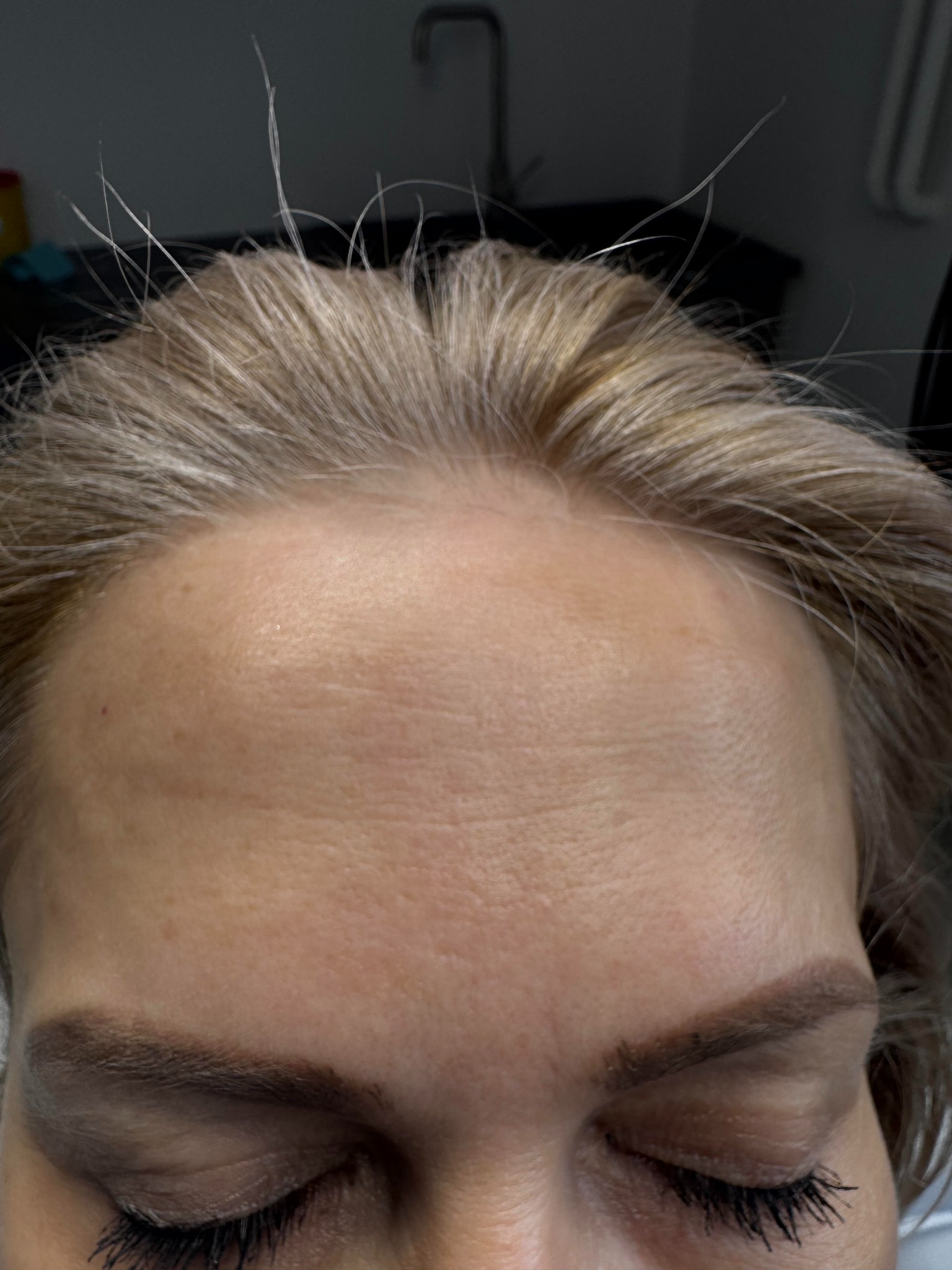 Zbliżenie na czoło i brwi kobiety, widoczne pojedyncze, odstające włosy na czubku głowy, skóra z widocznymi porami i zmarszczkami mimicznymi, ciemne brwi i rzęsy podkreślone tuszem.