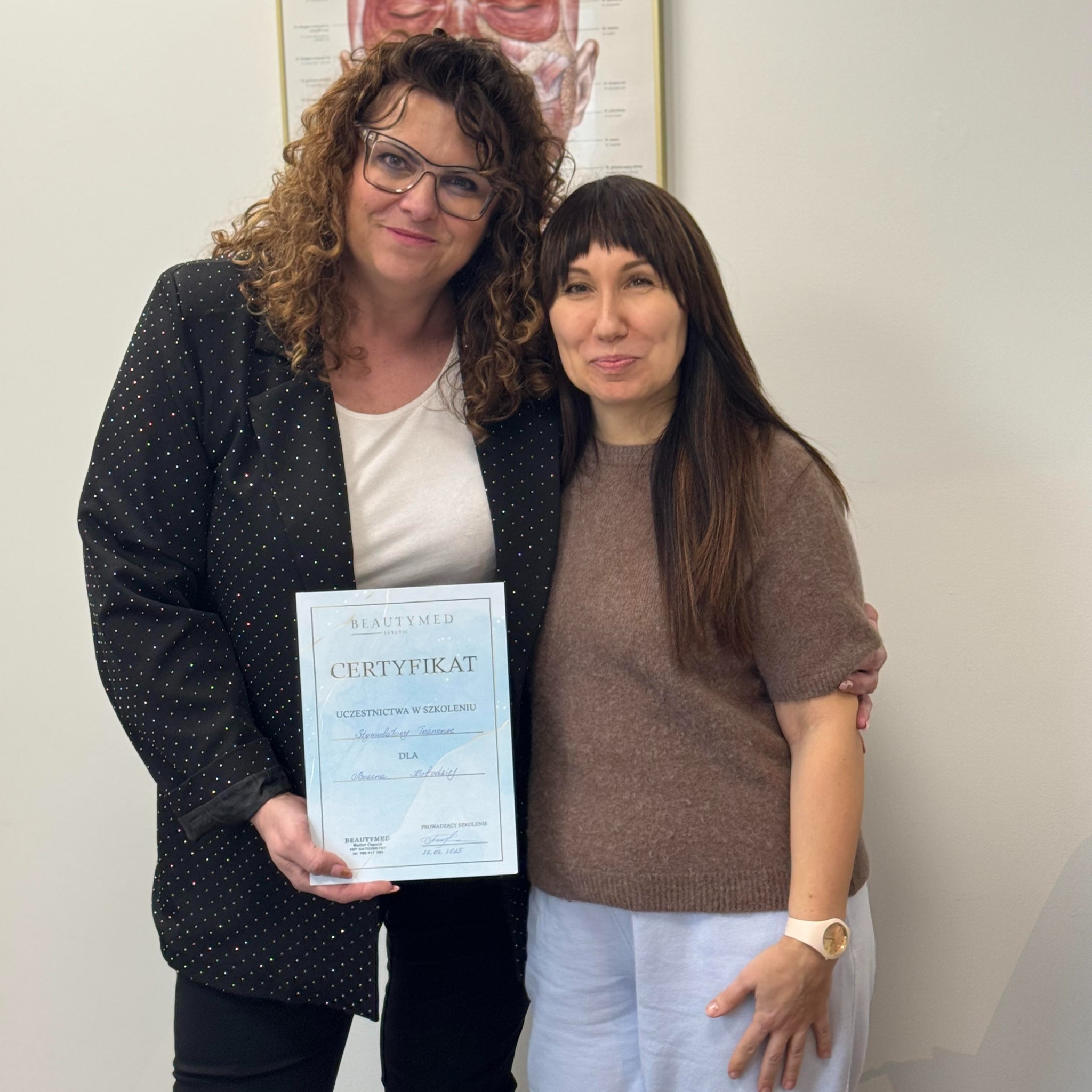 Dwie kobiety pozują z certyfikatem ukończenia szkolenia 'Stymulatory tkankowe' marki Beautymed, na tle ściany z plakatem anatomicznym.