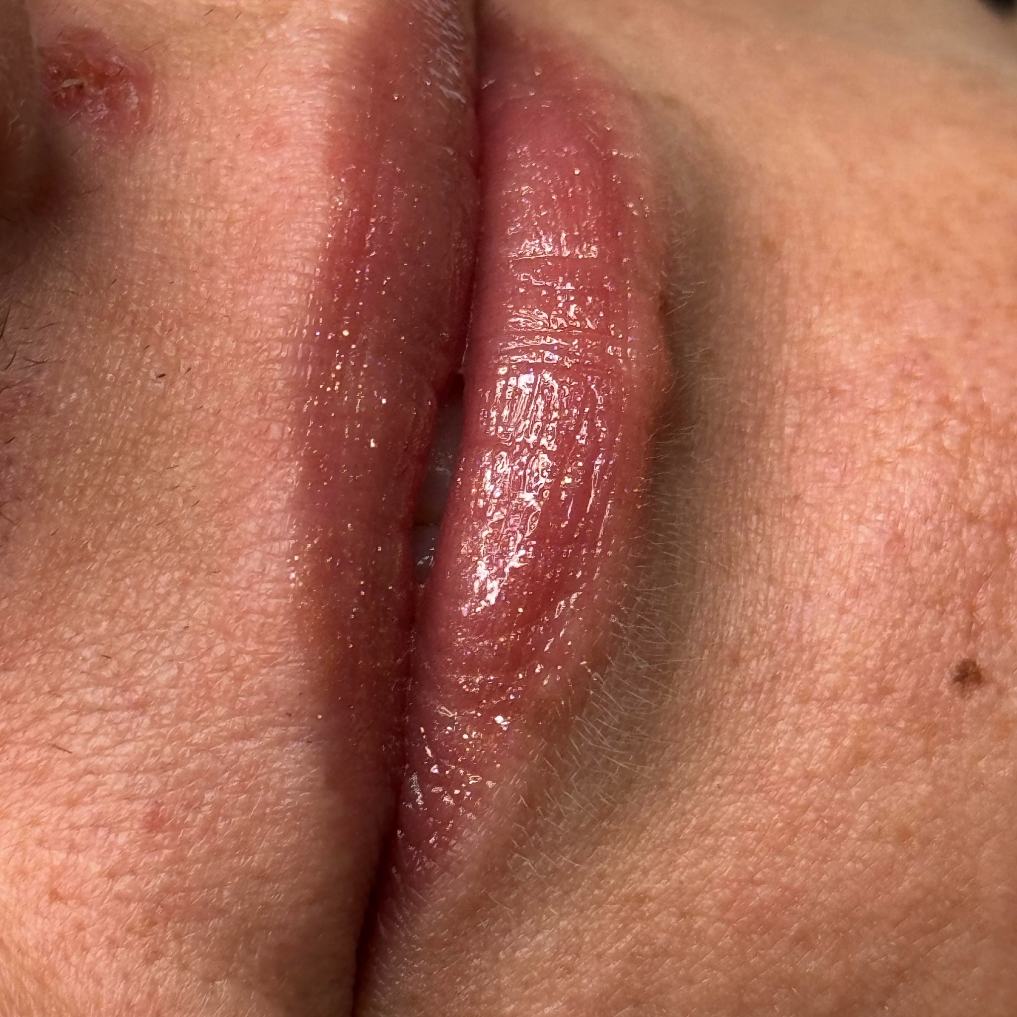 Powiększony widok ust z nałożonym błyszczykiem, widoczne drobne niedoskonałości skóry i włoski na twarzy.