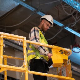 GRUPA SAFETOM SPÓŁKA Z OGRANICZONĄ ODPOWIEDZIALNOŚCIĄ - Pracownik w kasku i kamizelce odblaskowej na podnośniku koszowym podczas prac instalacyjnych pod sufitem, widoczne kable i elementy konstrukcyjne.