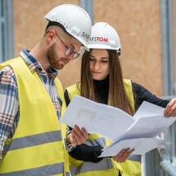 GRUPA SAFETOM SPÓŁKA Z OGRANICZONĄ ODPOWIEDZIALNOŚCIĄ - Dwoje pracowników w kamizelkach odblaskowych i kaskach z logo SAFE-TOM analizuje plany na placu budowy.