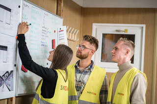 Trzy osoby w kamizelkach odblaskowych analizują schemat na tablicy podczas szkolenia, kobieta zaznacza element pisakiem.
