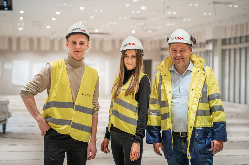 Trzy osoby w odzieży roboczej i kaskach z logo SAFETOM, stojące w jasnym, przestronnym wnętrzu w trakcie realizacji inwestycji.