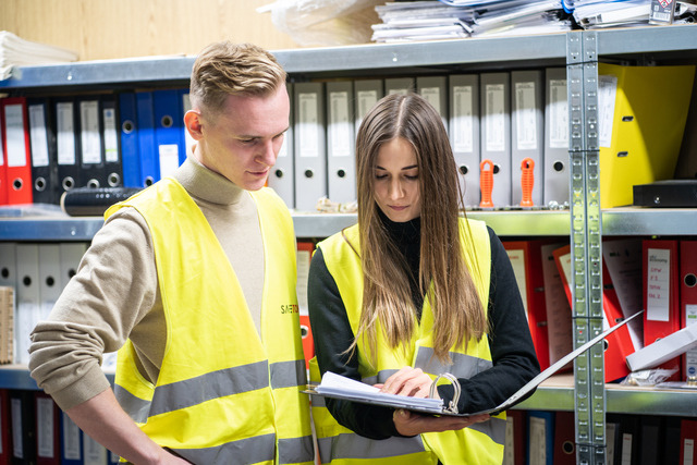 Dwoje pracowników w żółtych kamizelkach odblaskowych przegląda dokumenty w magazynie pełnym segregatorów.