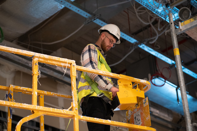 Pracownik w kasku i kamizelce odblaskowej na podnośniku koszowym podczas prac instalacyjnych pod sufitem, widoczne kable i elementy konstrukcyjne.