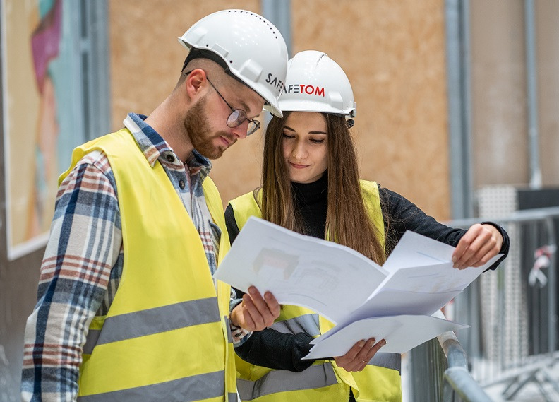 Dwoje pracowników w kamizelkach odblaskowych i kaskach z logo SAFE-TOM analizuje plany na placu budowy.