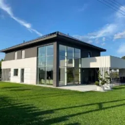 Nowoczesny dom dwukondygnacyjny z dużymi przeszkleniami i płaskim dachem, wykończony w bieli i szarości, otoczony zadbanym trawnikiem i zielenią.