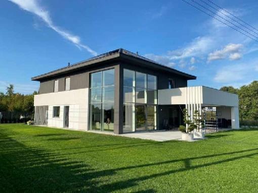 Nowoczesny dom dwukondygnacyjny z dużymi przeszkleniami i płaskim dachem, wykończony w bieli i szarości, otoczony zadbanym trawnikiem i zielenią.