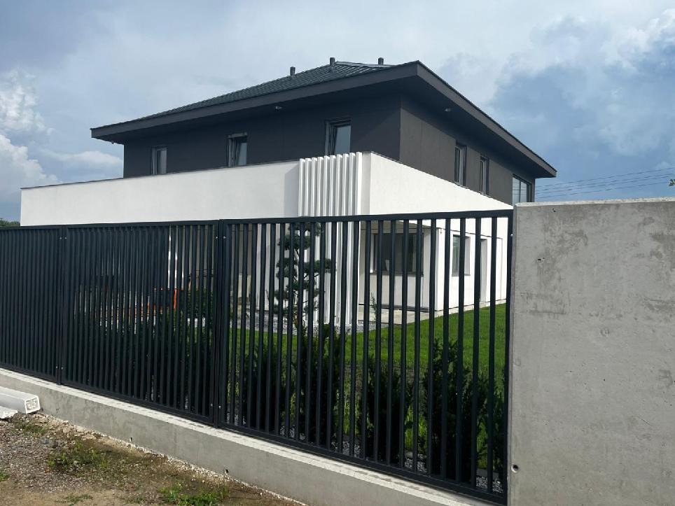 Nowoczesny dom dwukondygnacyjny z płaskim dachem, wykończony w kolorach szarości i bieli, z metalowym ogrodzeniem panelowym i betonowym murem.