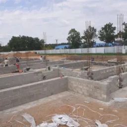 Wznoszenie fundamentów domu z bloczków betonowych, widoczne zbrojenia pionowe, pracownicy budowlani w tle, teren budowy w trakcie prac.