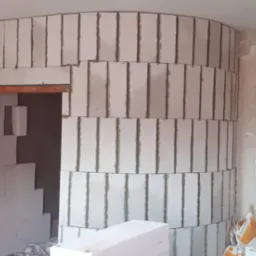 Wnętrze w trakcie prac murarskich: półokrągła ściana z białych bloczków, widoczne spoiny, fragment otworu drzwiowego z nadprożem, pracownik w tle oraz worki z materiałami budowlanymi.