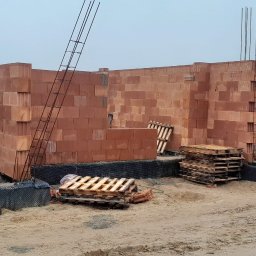 Damian Kuś Usługi Budowlane - Wznoszenie ścian z czerwonej ceramiki poryzowanej na placu budowy. Widoczne stalowe pręty zbrojeniowe, palety, oraz folia fundamentowa. Szaro-błękitne niebo w tle.