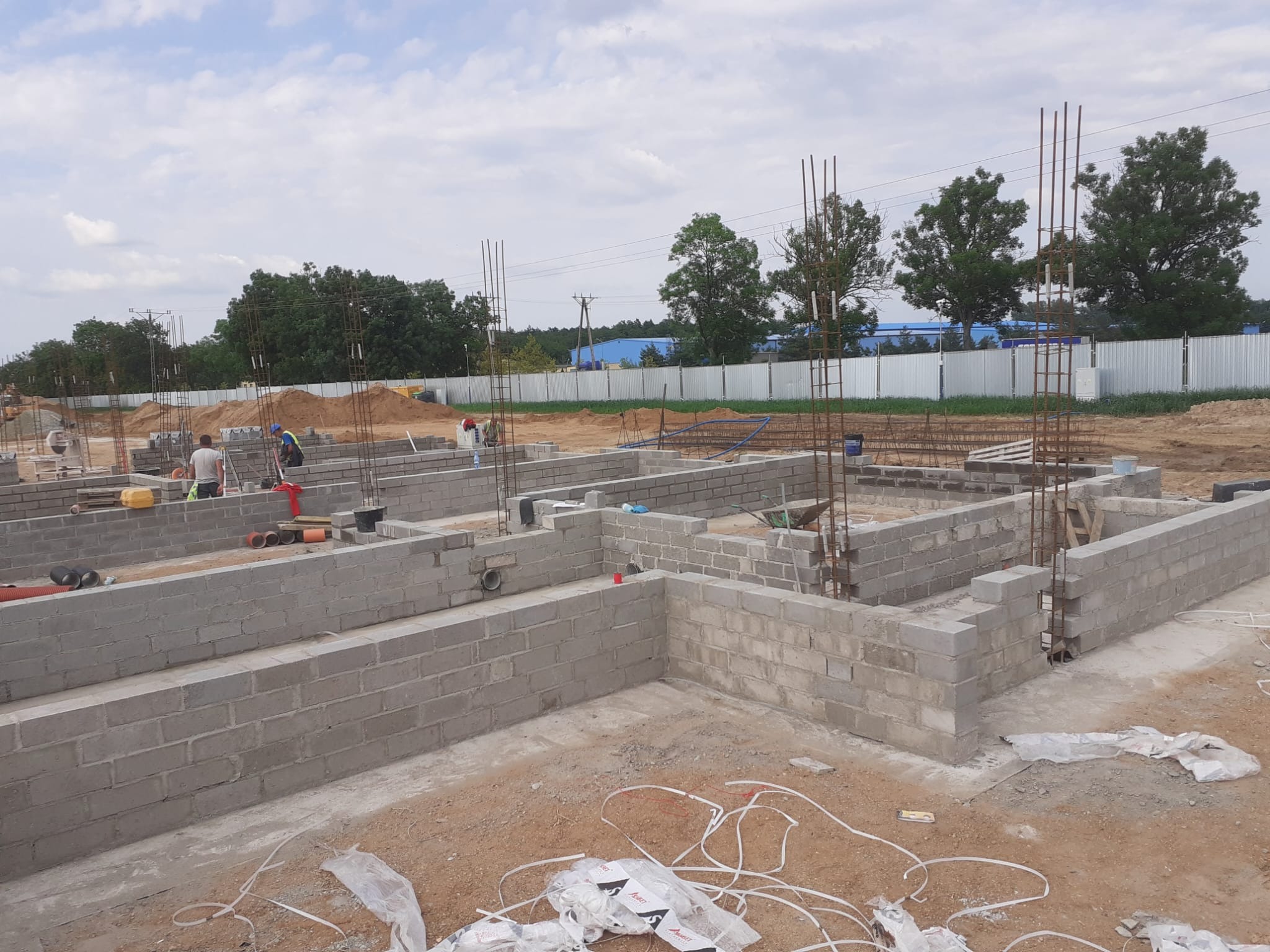 Wznoszenie fundamentów domu z bloczków betonowych, widoczne zbrojenia pionowe, pracownicy budowlani w tle, teren budowy w trakcie prac.