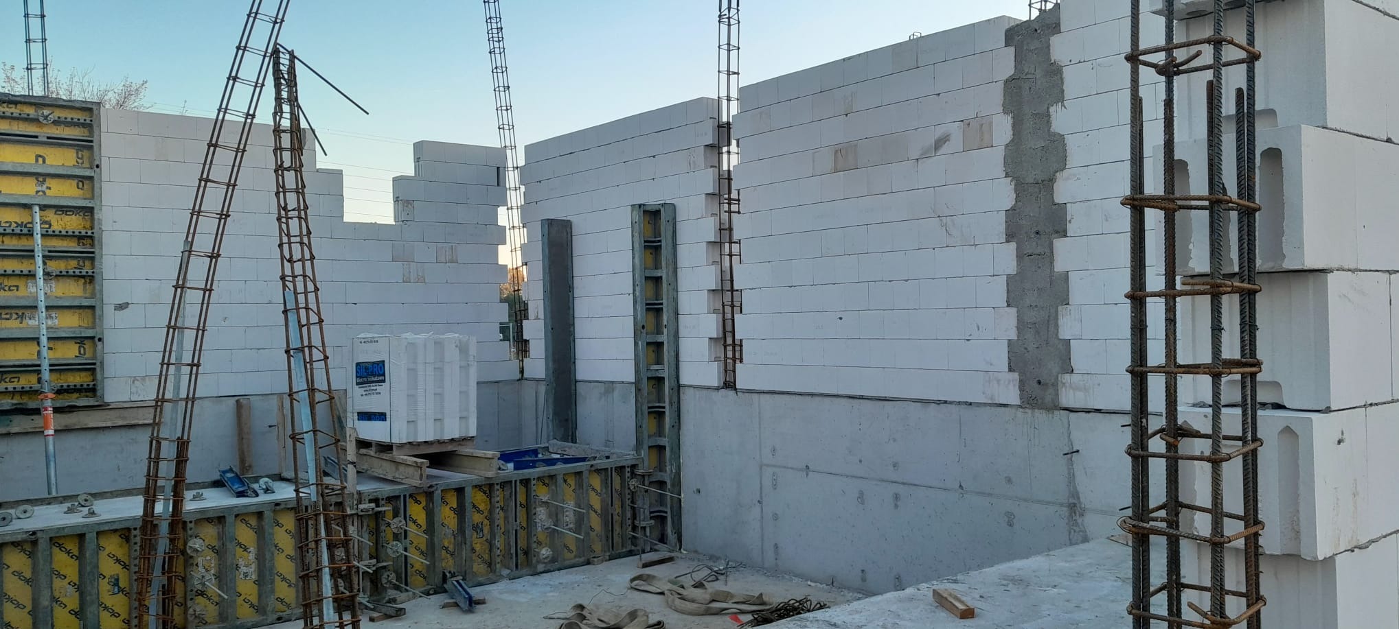 Wznoszenie ścian z białych bloczków betonowych na fundamencie zbrojonym stalą, widoczne szalunki i elementy konstrukcyjne.