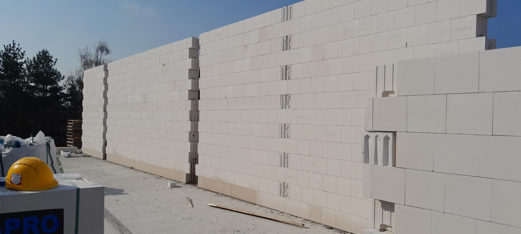 Wznoszone ściany z białych bloczków betonu komórkowego, widoczne spoiny pionowe oraz żółty kask ochronny i paleta bloczków w tle.