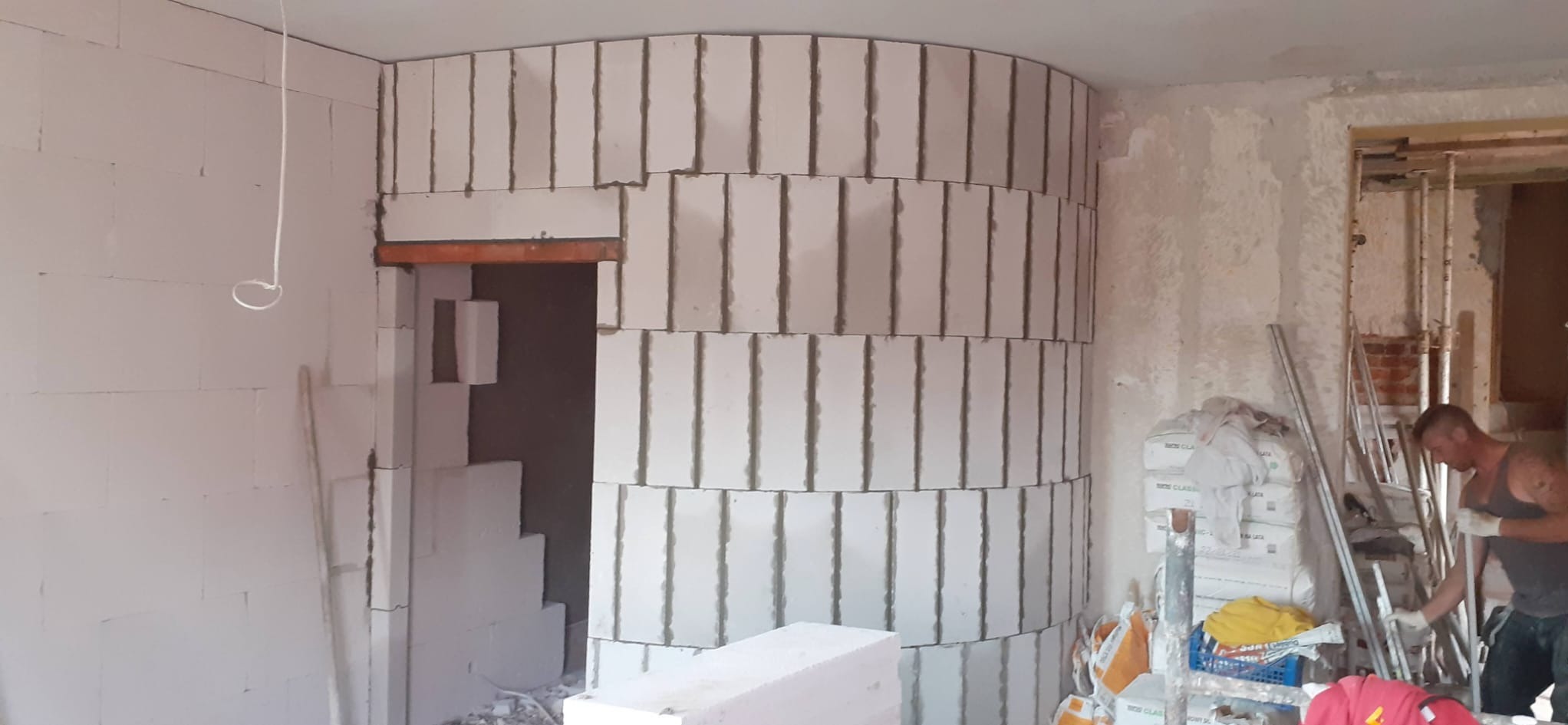 Wnętrze w trakcie prac murarskich: półokrągła ściana z białych bloczków, widoczne spoiny, fragment otworu drzwiowego z nadprożem, pracownik w tle oraz worki z materiałami budowlanymi.