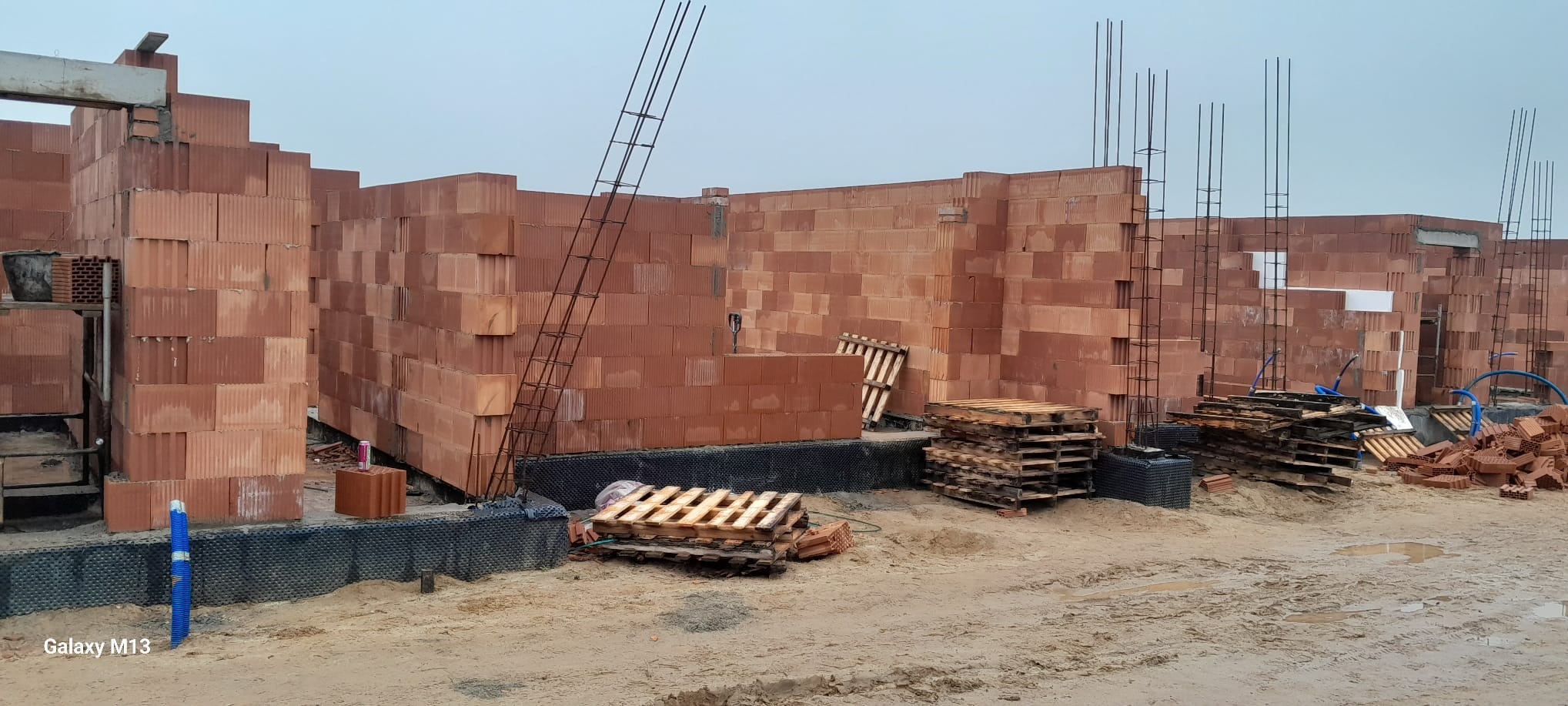 Wznoszenie ścian z czerwonej ceramiki poryzowanej na placu budowy. Widoczne stalowe pręty zbrojeniowe, palety, oraz folia fundamentowa. Szaro-błękitne niebo w tle.