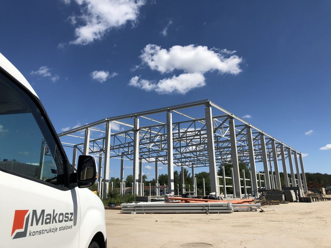 Wznoszona konstrukcja stalowa hali, widok z boku samochodu firmowego z logo 'Makosz konstrukcje stalowe', błękitne niebo z chmurami w tle.