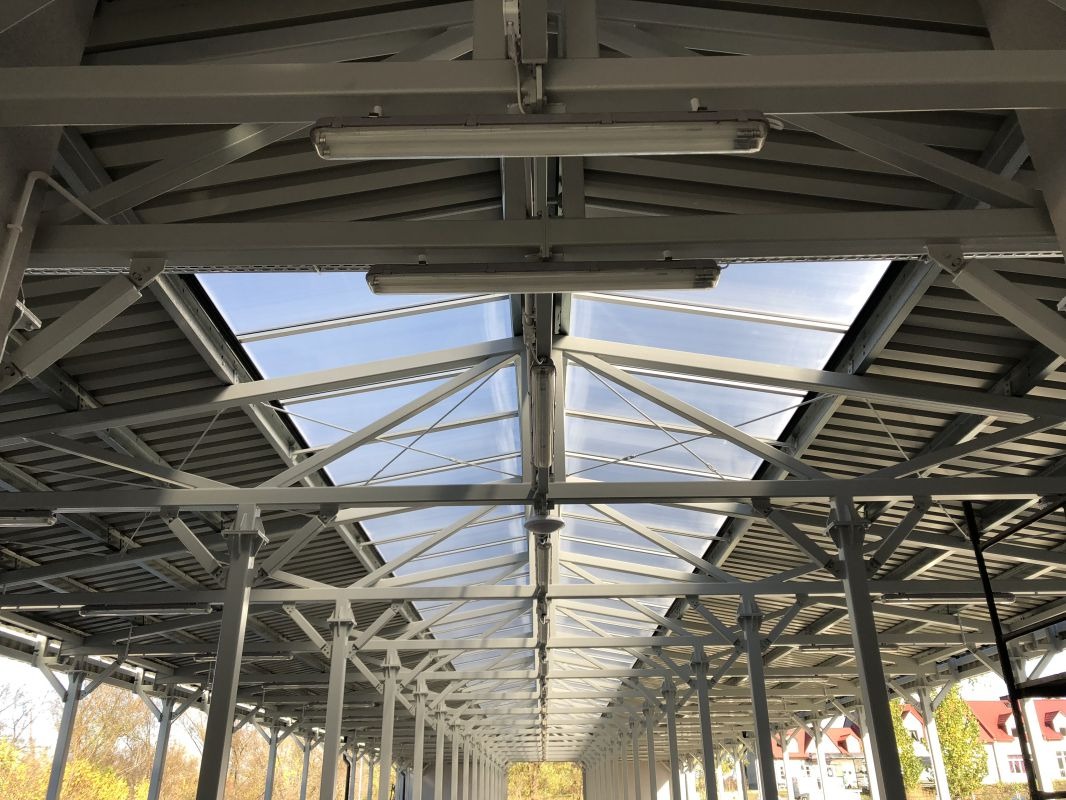 Wnętrze hali z widoczną stalową konstrukcją dachu i oświetleniem, z przeszkleniami w dachu przepuszczającymi światło dzienne.