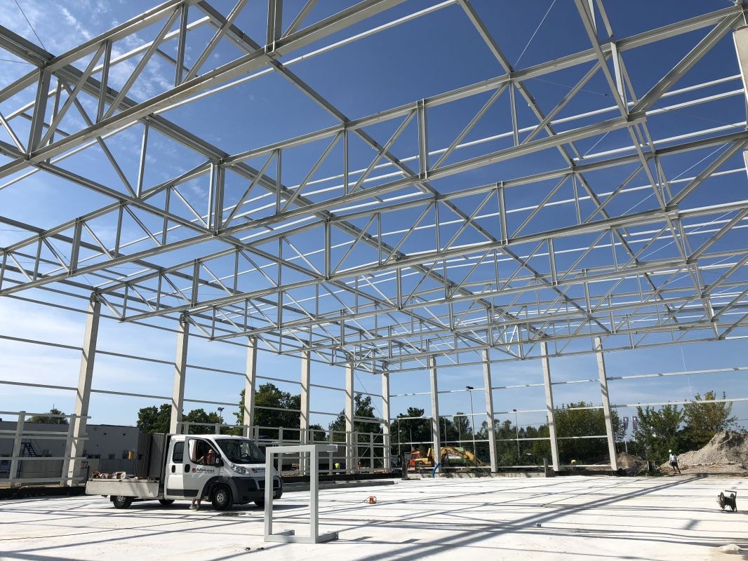 Wnętrze budowanej hali z widoczną stalową konstrukcją dachu i betonowymi słupami, na zewnątrz zaparkowany biały samochód dostawczy z materiałami budowlanymi, w tle zieleń drzew i błękitne niebo.