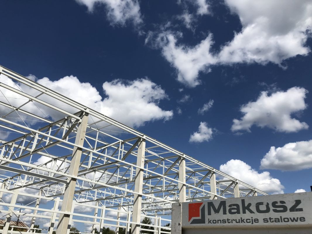 Konstrukcja stalowa hali w trakcie budowy, widoczne białe elementy szkieletu na tle błękitnego nieba z chmurami, w dolnej części logo firmy Makosz Konstrukcje Stalowe.