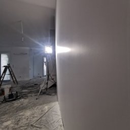 APEX Agregat Painting Expert - Świeżo wyszpachlowana ściana w pomieszczeniu w trakcie prac wykończeniowych, widoczny reflektor na statywie oświetlający powierzchnię, podłoga zabezpieczona folią malarską, drabina i kable w tle.