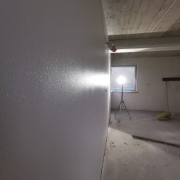 APEX Agregat Painting Expert - Świeżo pomalowana ściana w szarym kolorze, widoczna faktura, w tle lampa budowlana na statywie oświetlająca pomieszczenie w trakcie prac.