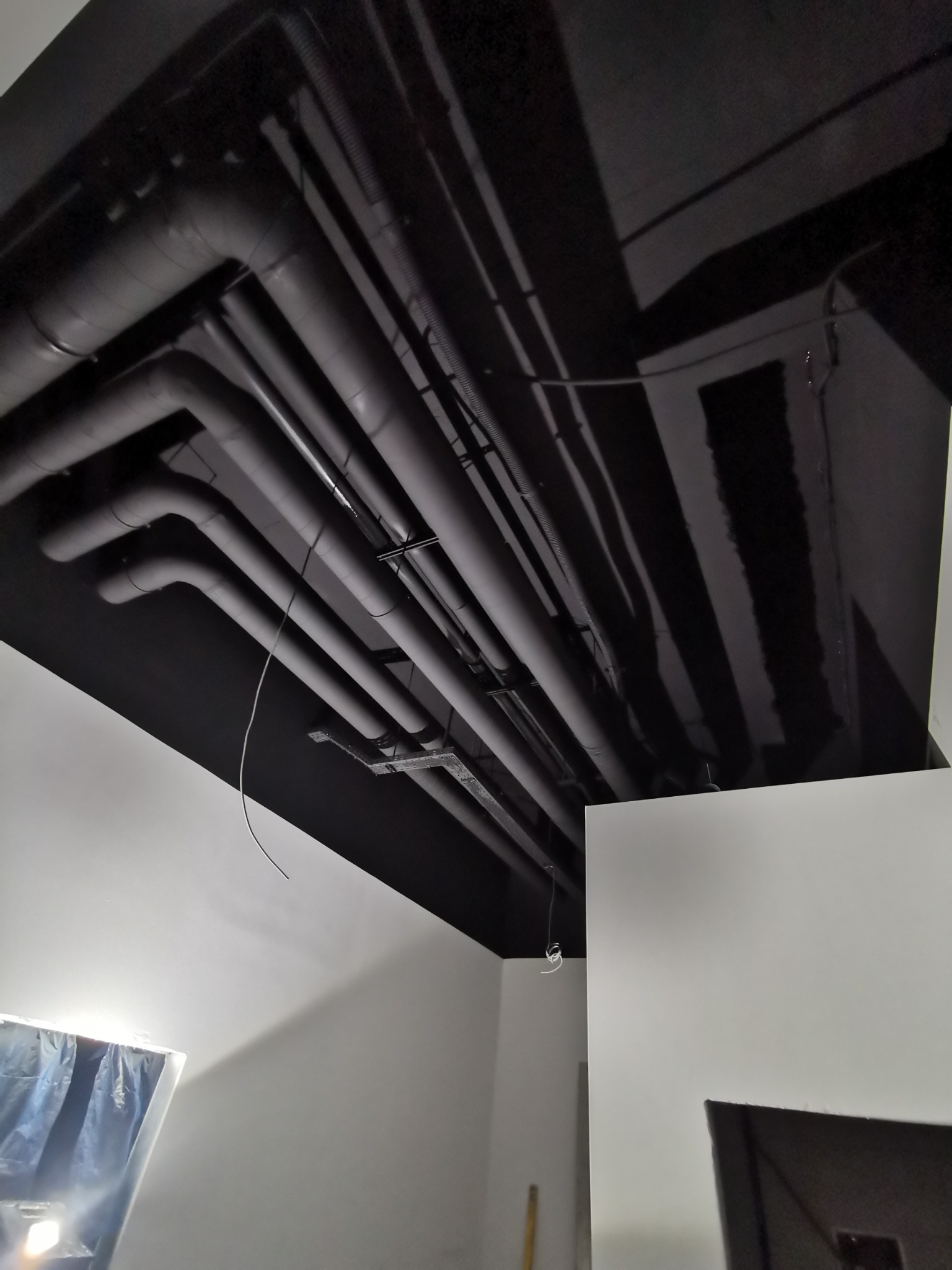 Czarny sufit z widocznymi rurami pomalowanymi na czarno, białe ściany w pomieszczeniu w trakcie prac wykończeniowych.