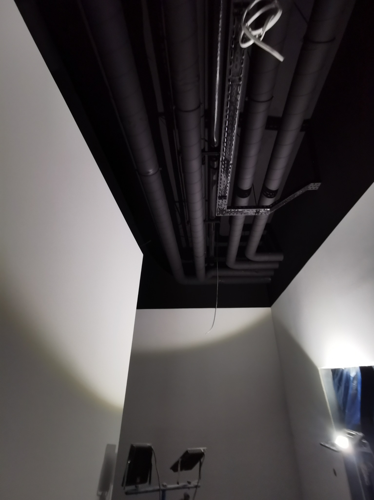 Czarny sufit z widocznymi rurami wentylacyjnymi i metalowymi korytkami kablowymi, oświetlenie robocze w pomieszczeniu z białymi ścianami.