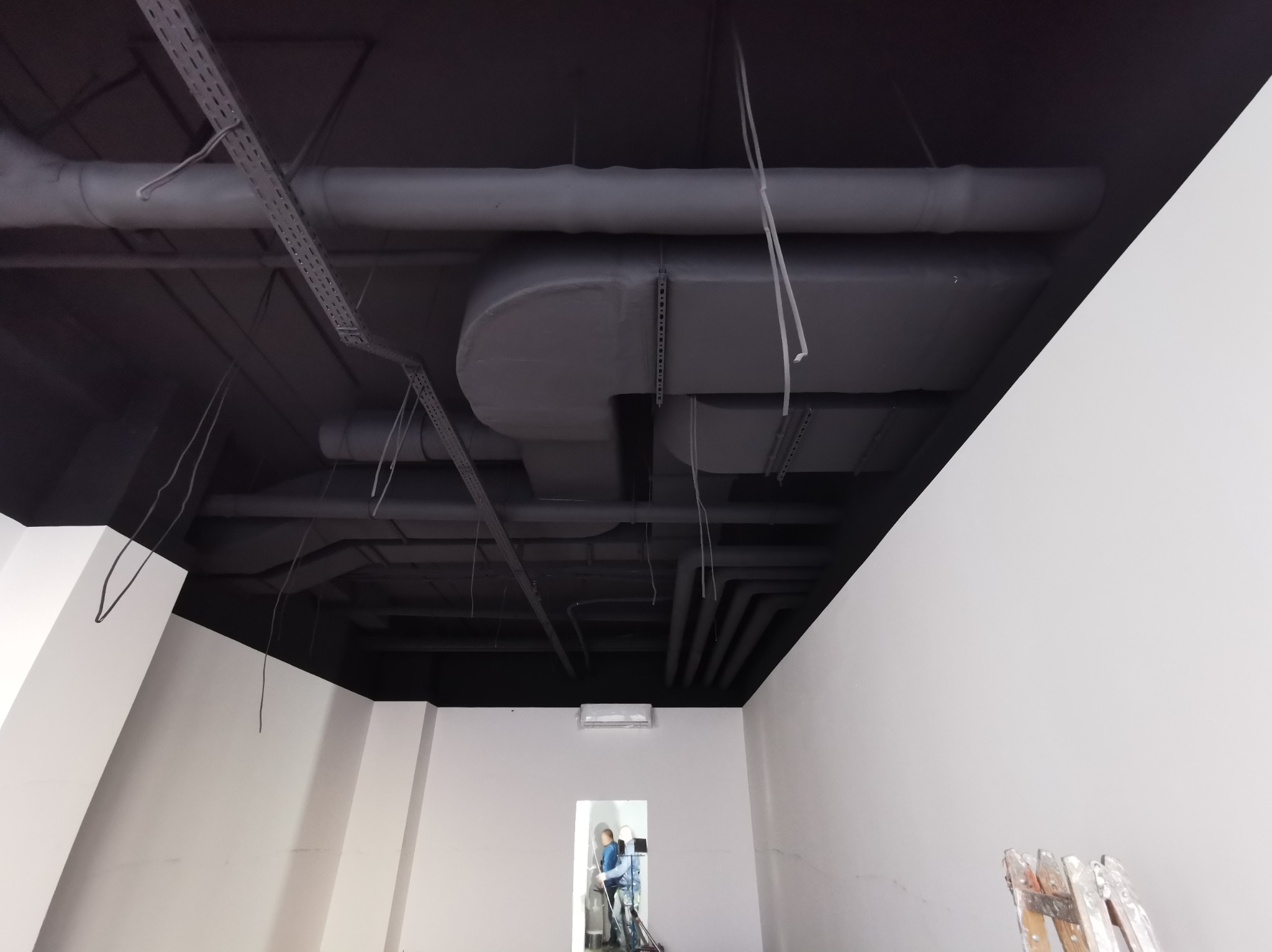 Pomieszczenie w trakcie prac wykończeniowych: pomalowany na czarno sufit z widocznymi rurami i instalacjami, białe ściany, drabina w rogu i pracownik w oddali.