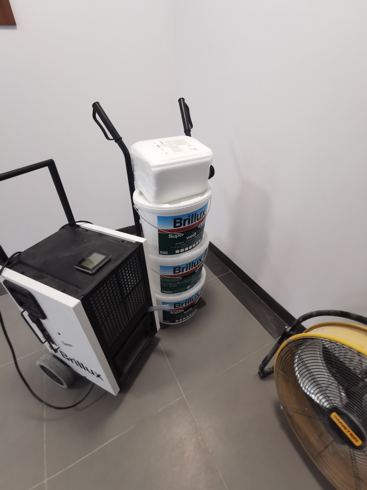 Stos puszek z farbą marki Brillux na wózku obok osuszacza powietrza i wentylatora na szarej podłodze w pomieszczeniu ze ścianami w odcieniach bieli.