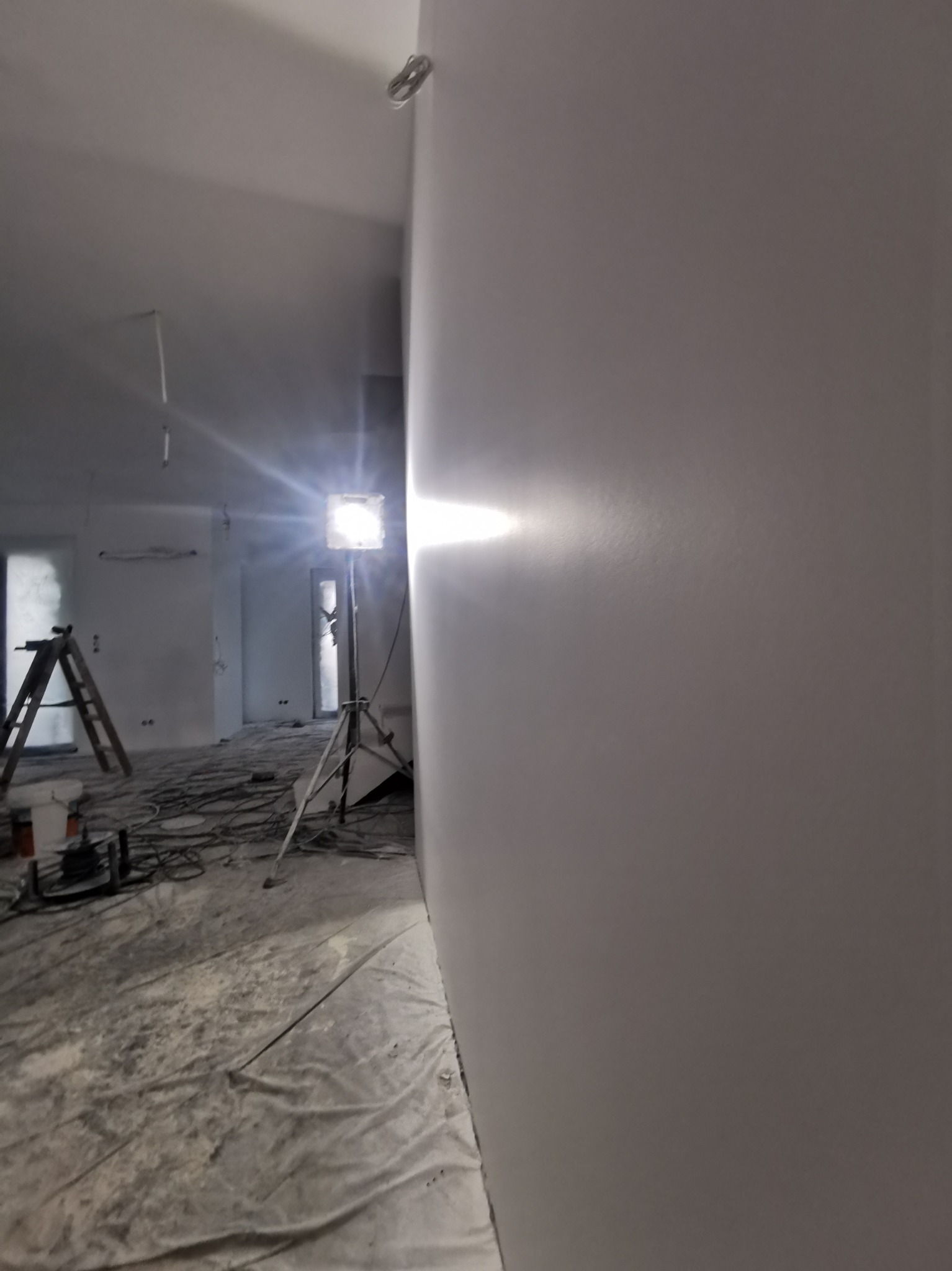 Świeżo wyszpachlowana ściana w pomieszczeniu w trakcie prac wykończeniowych, widoczny reflektor na statywie oświetlający powierzchnię, podłoga zabezpieczona folią malarską, drabina i kable w tle.