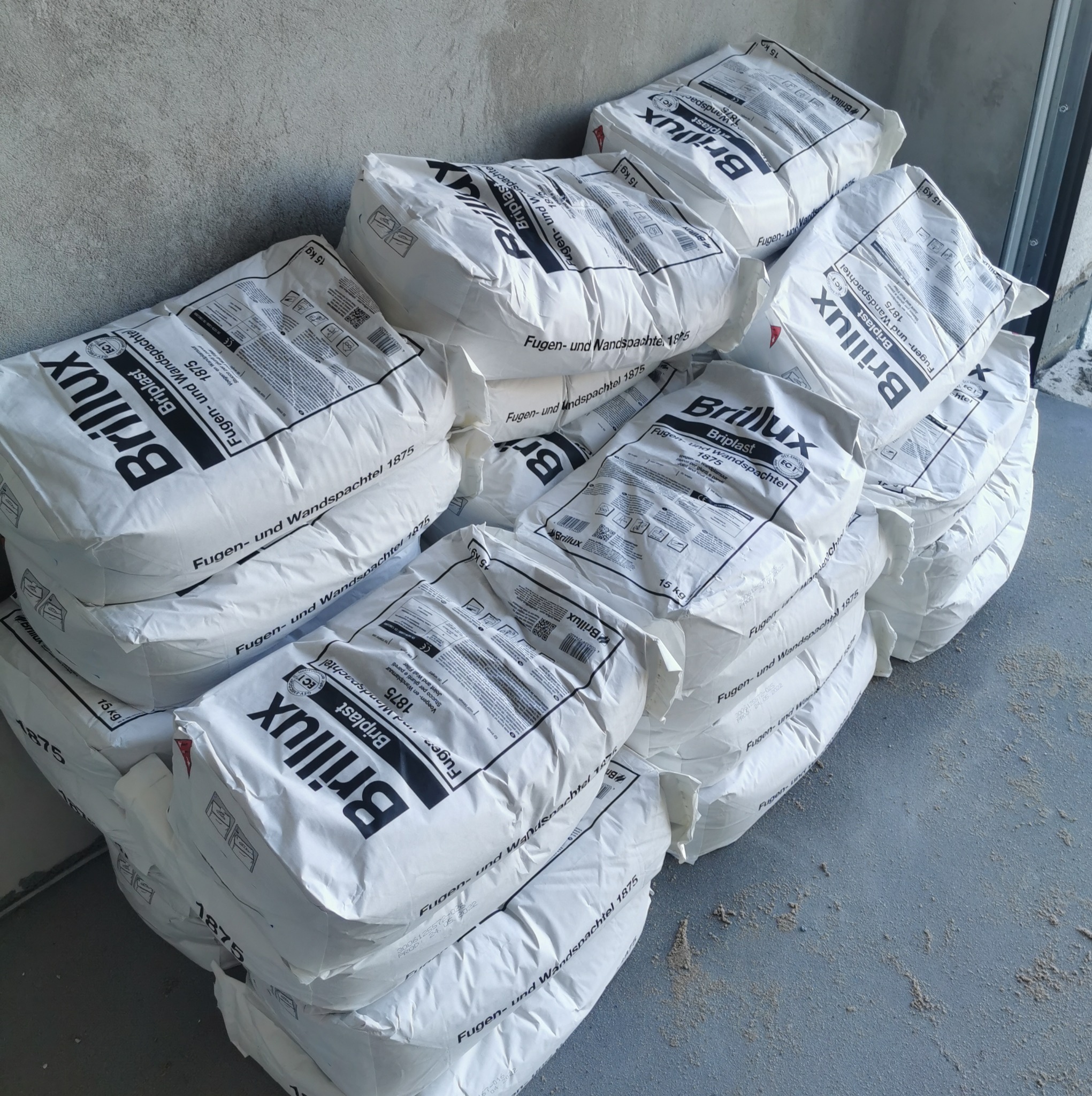 Stos białych worków 15kg z zaprawą szpachlową Brillux Briplast 1875, ułożonych pod ścianą na szarej posadzce, widoczne etykiety z informacjami i logo producenta.