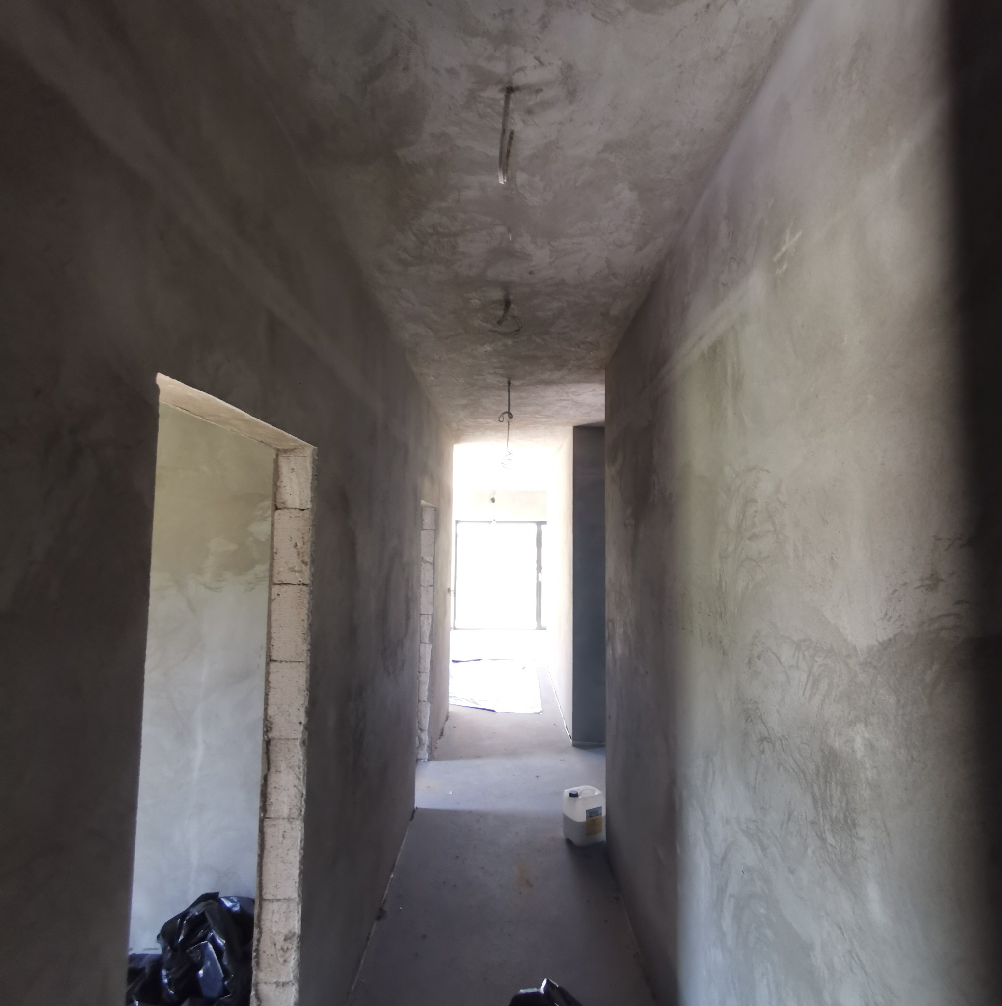 Korytarz w trakcie remontu, ściany i sufit pokryte szpachlą, widoczne przewody oświetleniowe, worek z odpadami budowlanymi i pojemnik z płynem do gruntowania.