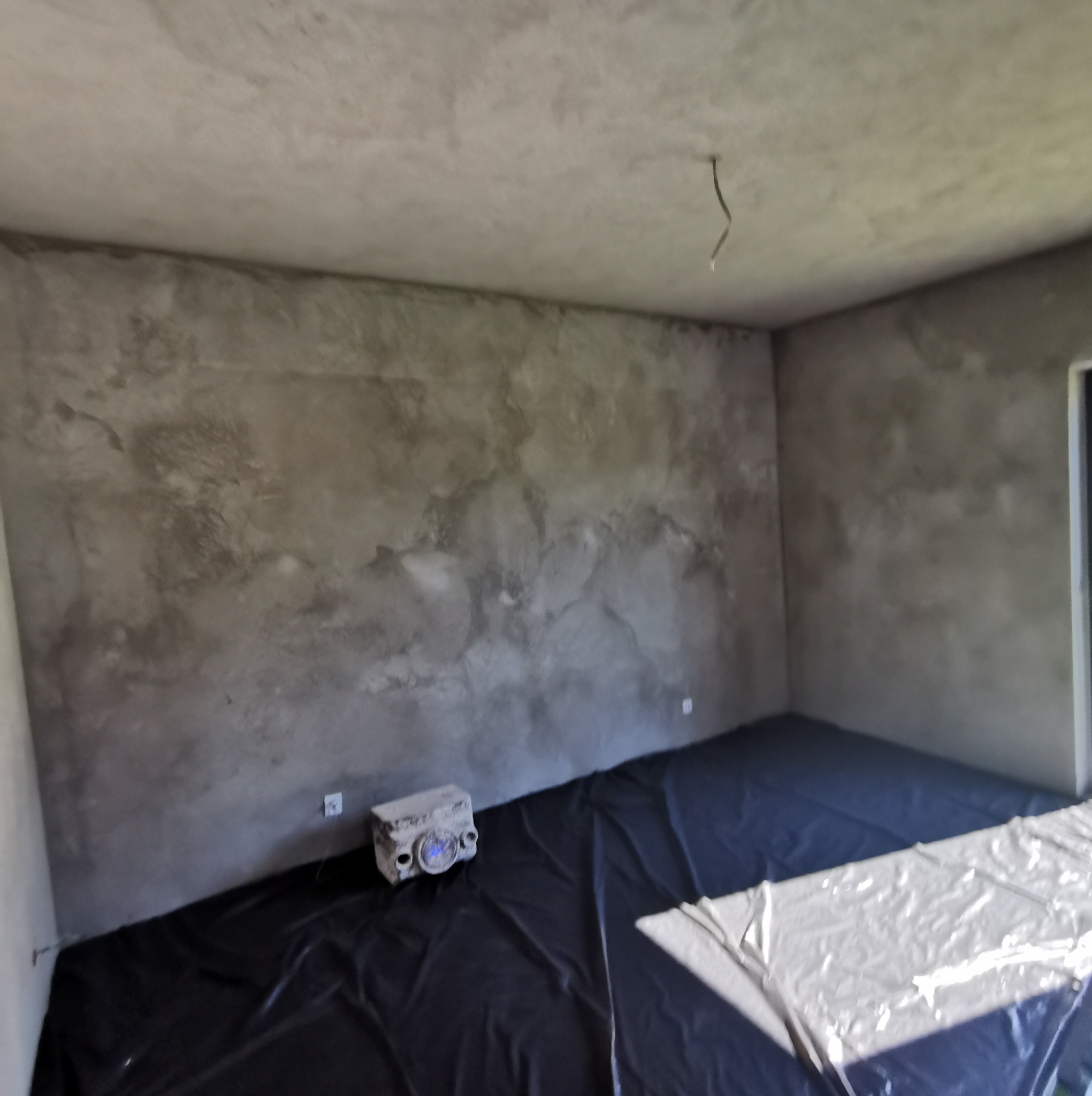 Surowe, szare ściany po nałożeniu wstępnej warstwy, podłoga zabezpieczona czarną folią, widoczna puszka elektryczna i wystający kabel ze stropu.