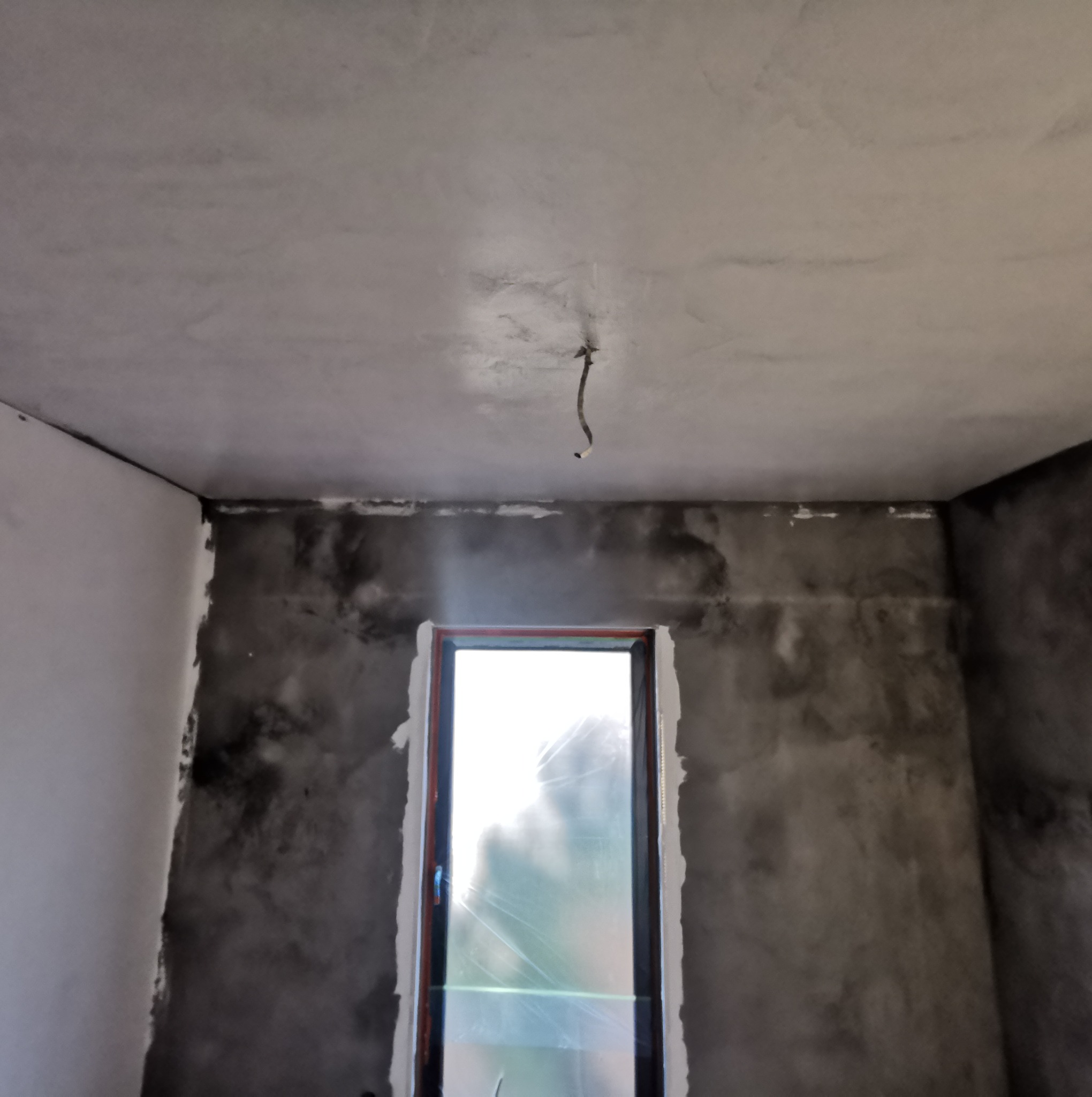 Pokój w trakcie prac wykończeniowych, ściany pokryte szarą, surową fakturą, okno zabezpieczone folią, widoczny fragment instalacji elektrycznej zwisający z sufitu.
