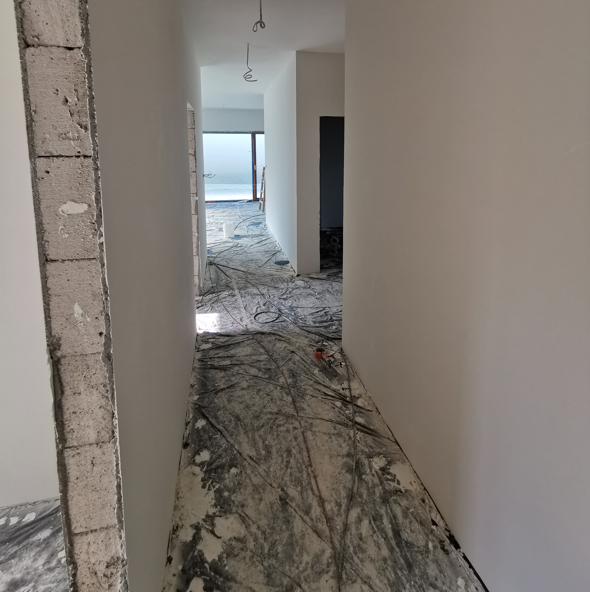 Korytarz w trakcie prac wykończeniowych, zabezpieczona podłoga folią malarską, ściany świeżo pokryte białą farbą, widoczne przewody elektryczne zwisające z sufitu i surowy, nieotynkowany fragment...